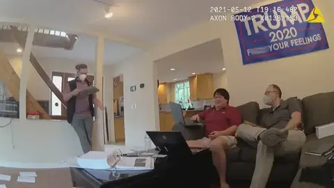 Police body-camera frame: James Zhong on his sofa, laptop open, showing investigators a wallet holding roughly 1,500 BTC while an agent takes notes